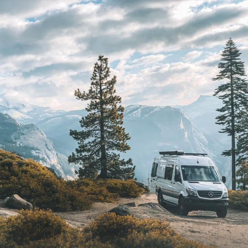 A Storyteller Overland van parked on a dirt path amidst pine trees and mountains under a cloudy sky, illustrating off-grid capability and outdoor exploration.