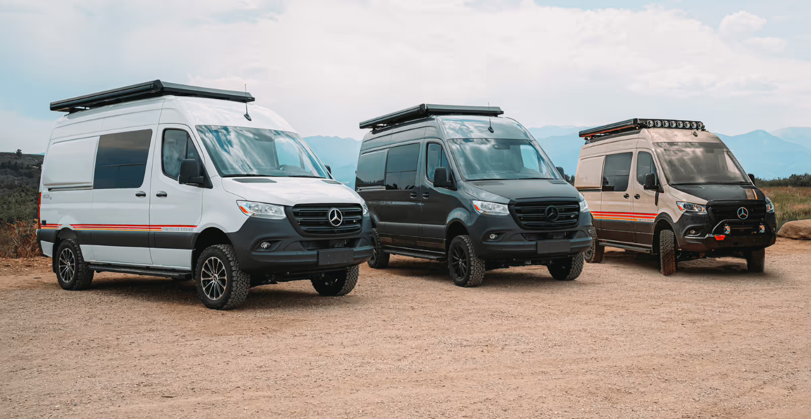 Three Storyteller Overland vans (Classic, Stealth, and Beast MODE) parked side-by-side in a vast outdoor landscape, representing the range of adventure vehicles offered by Mercedes-Benz of Birmingham.